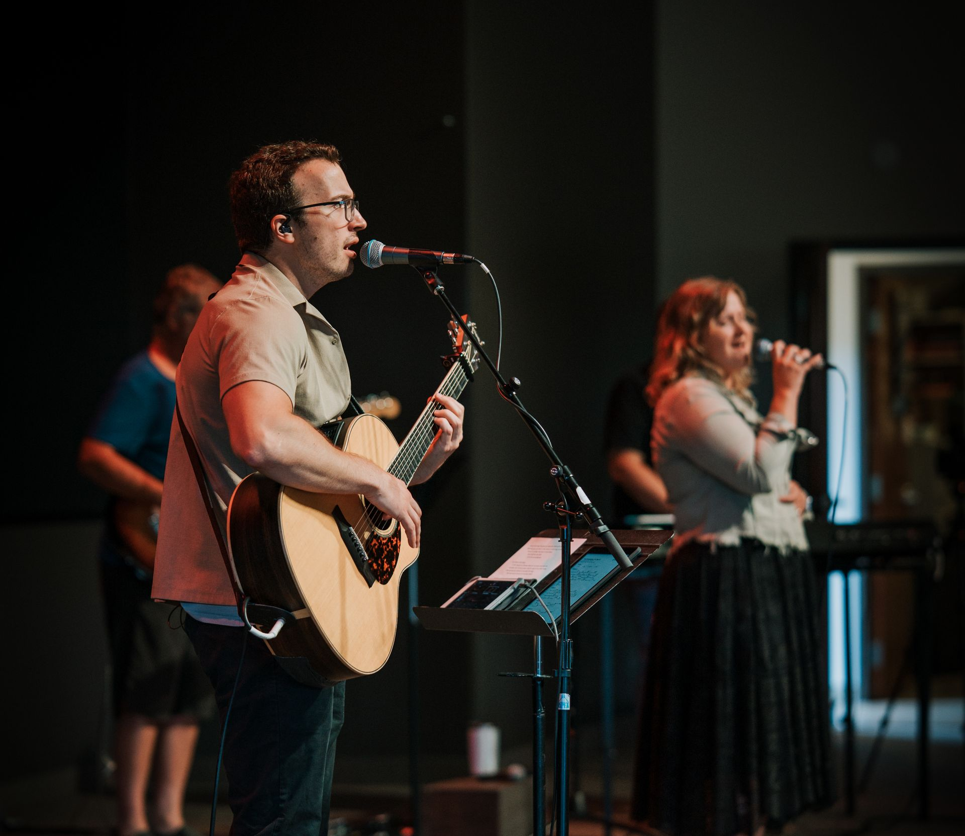 A band performs on stage: guitarist sings, female vocalist, other musicians in the background. Dim lighting.