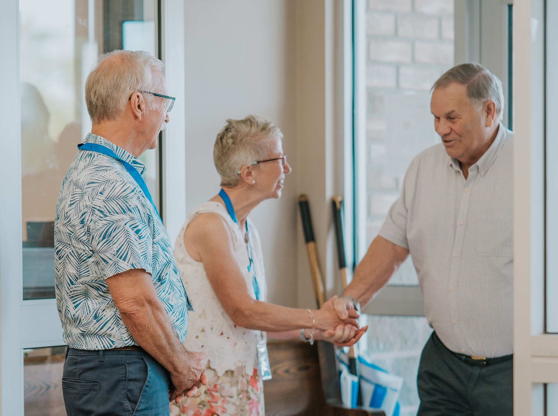Three people greet each other. Two people shake hands, inside a building with a glass door.