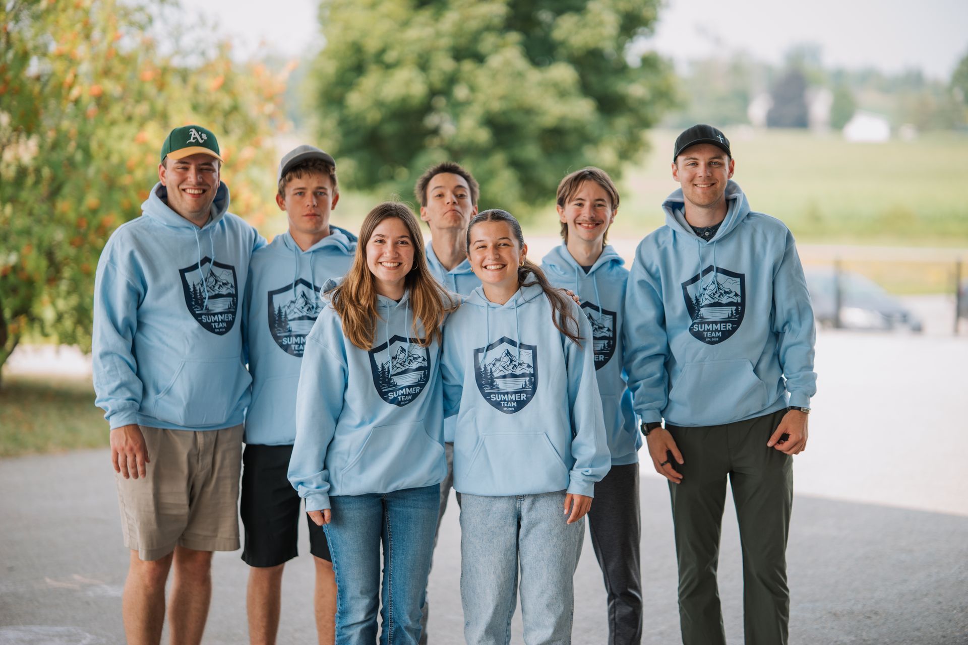 Group of people in light blue hoodies, smiling outdoors. Hoodies have a mountain logo.
