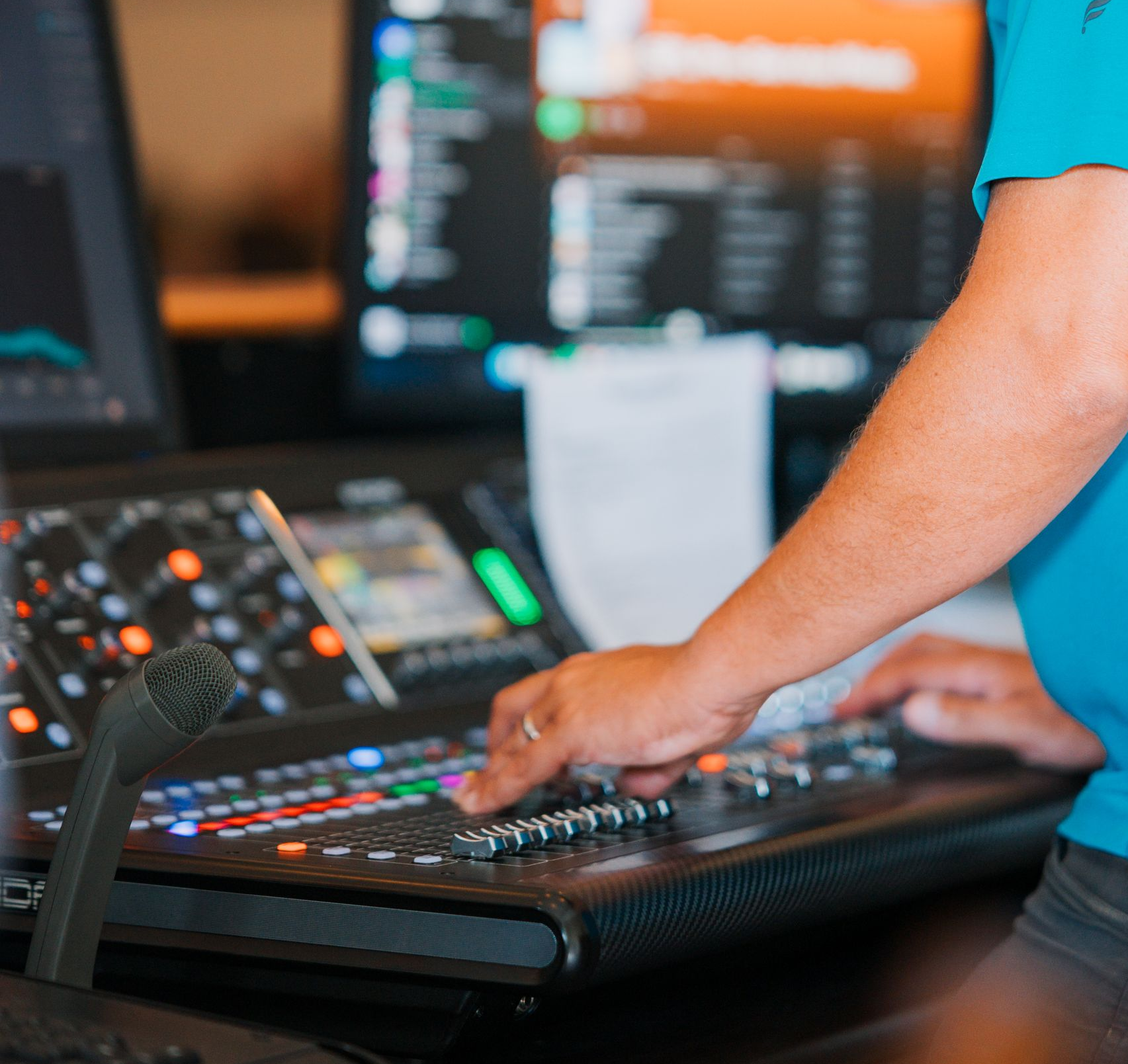 Person operates a sound mixing console with a microphone, multiple monitors in background.