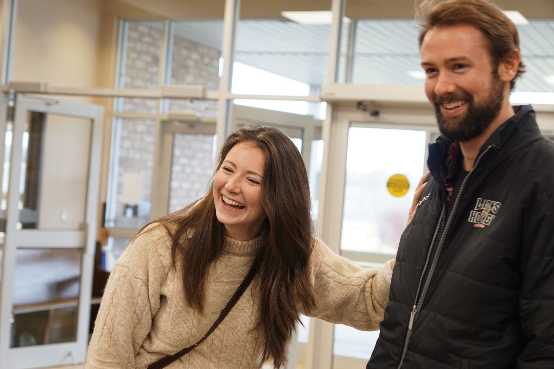 Woman laughing, arm on man's shoulder. They stand in a bright, interior space near a glass door.
