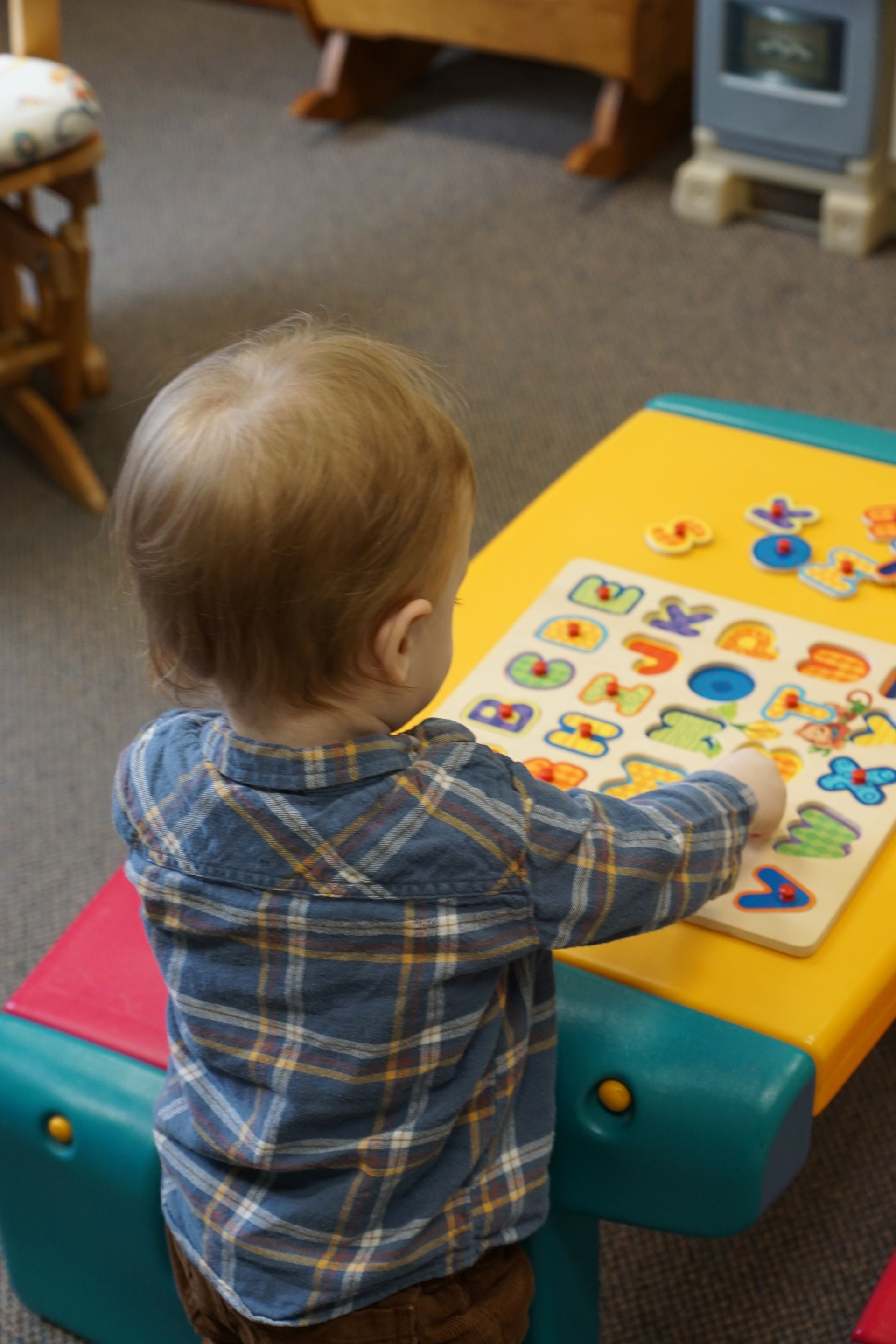 Child in plaid shirt working on an alphabet puzzle at a yellow table in a room.