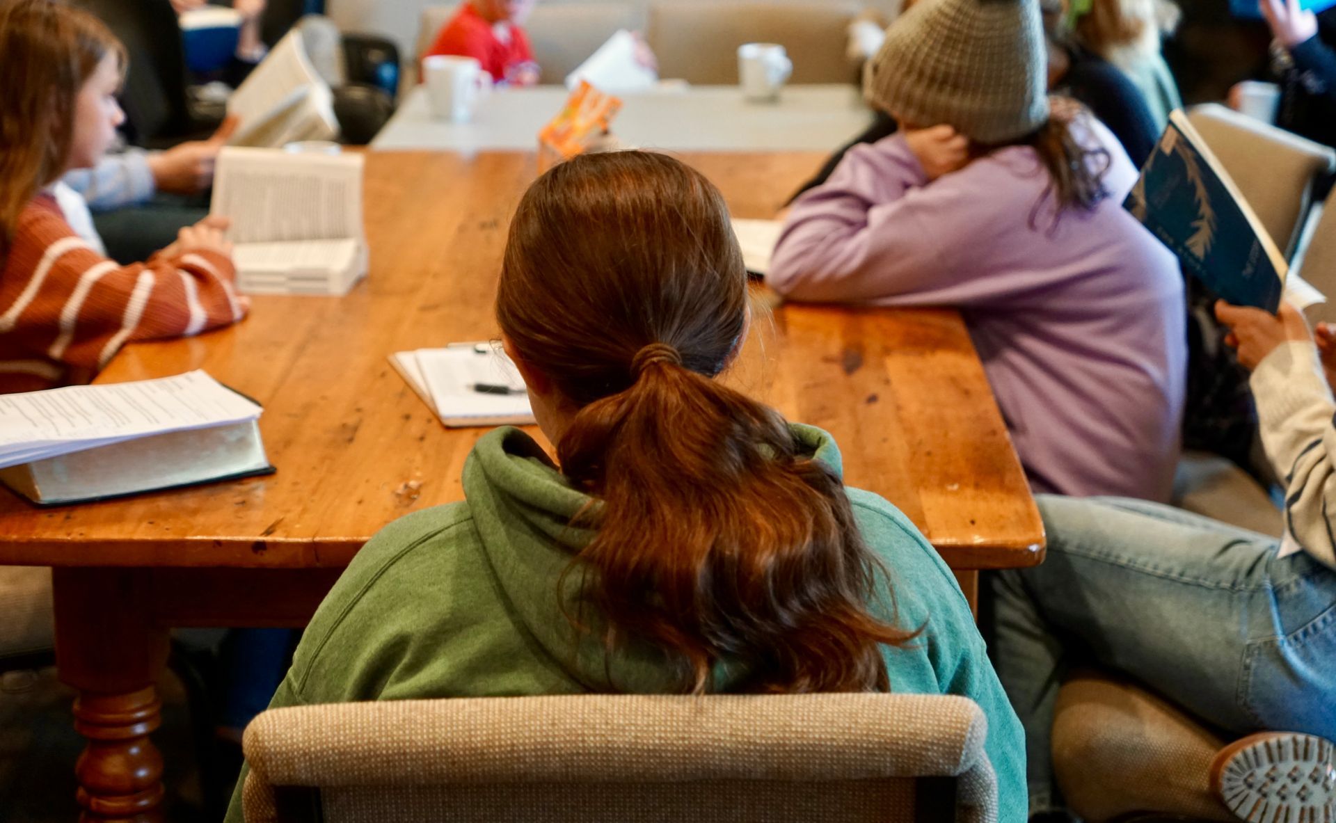 Group of people sitting at a wooden table, some with books, some in a casual setting.