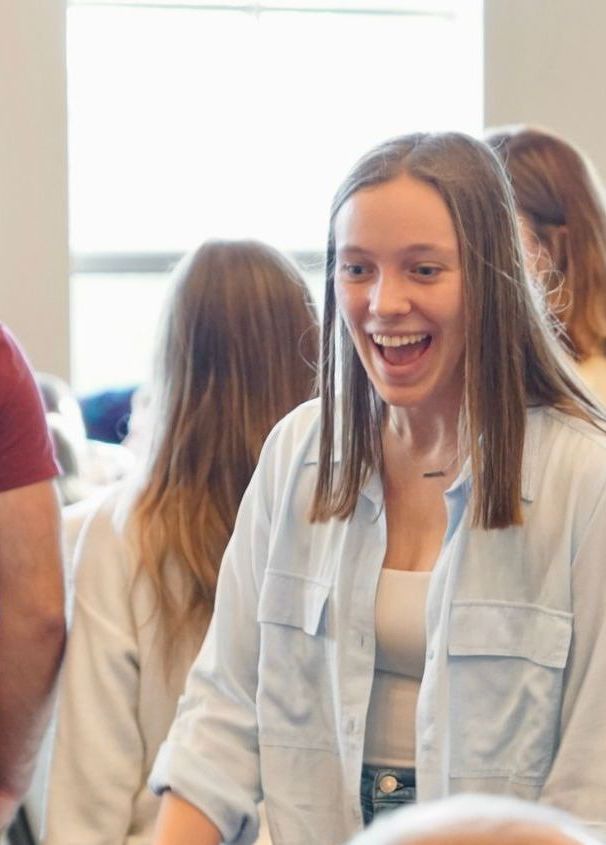 Woman with open mouth, smiling and looking to the side. Light blue button-down shirt, white top.