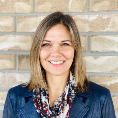 Woman smiles, wearing blue jacket and patterned scarf, against a brick wall.
