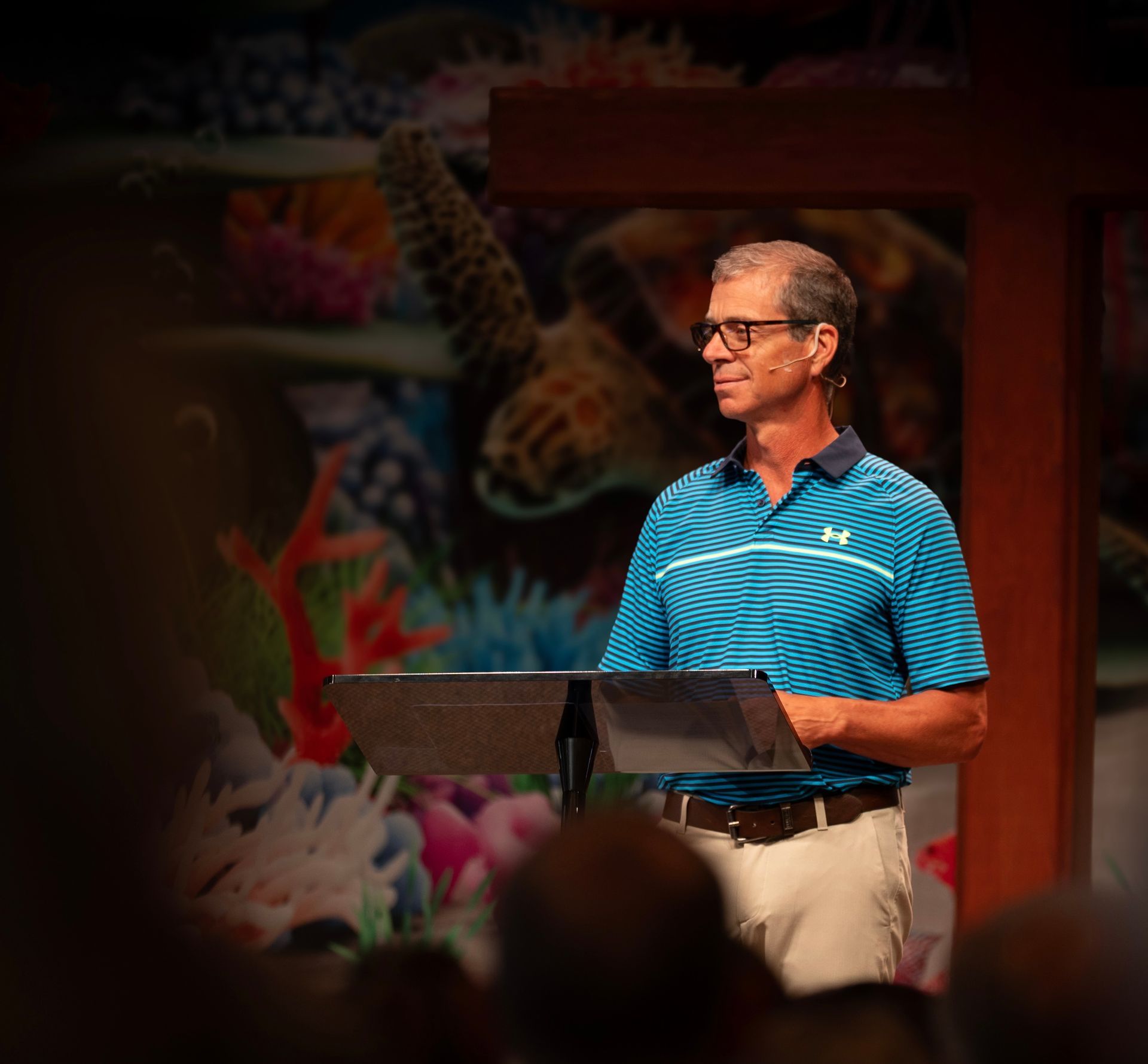 Man speaking at a lectern on stage, wearing a blue shirt. Backdrop features coral and a sea turtle.