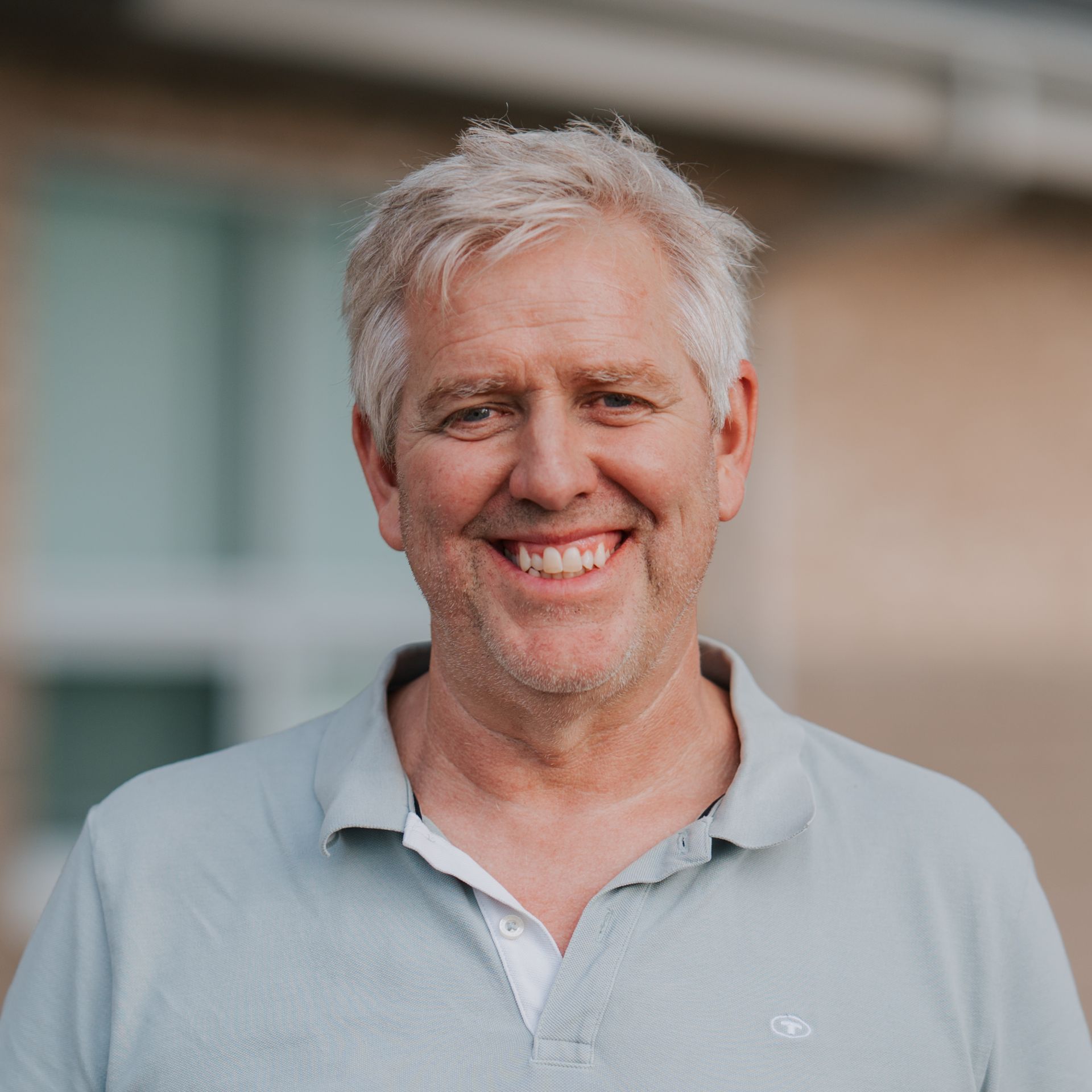 Man with gray hair smiling, wearing a light blue polo shirt, outdoors.