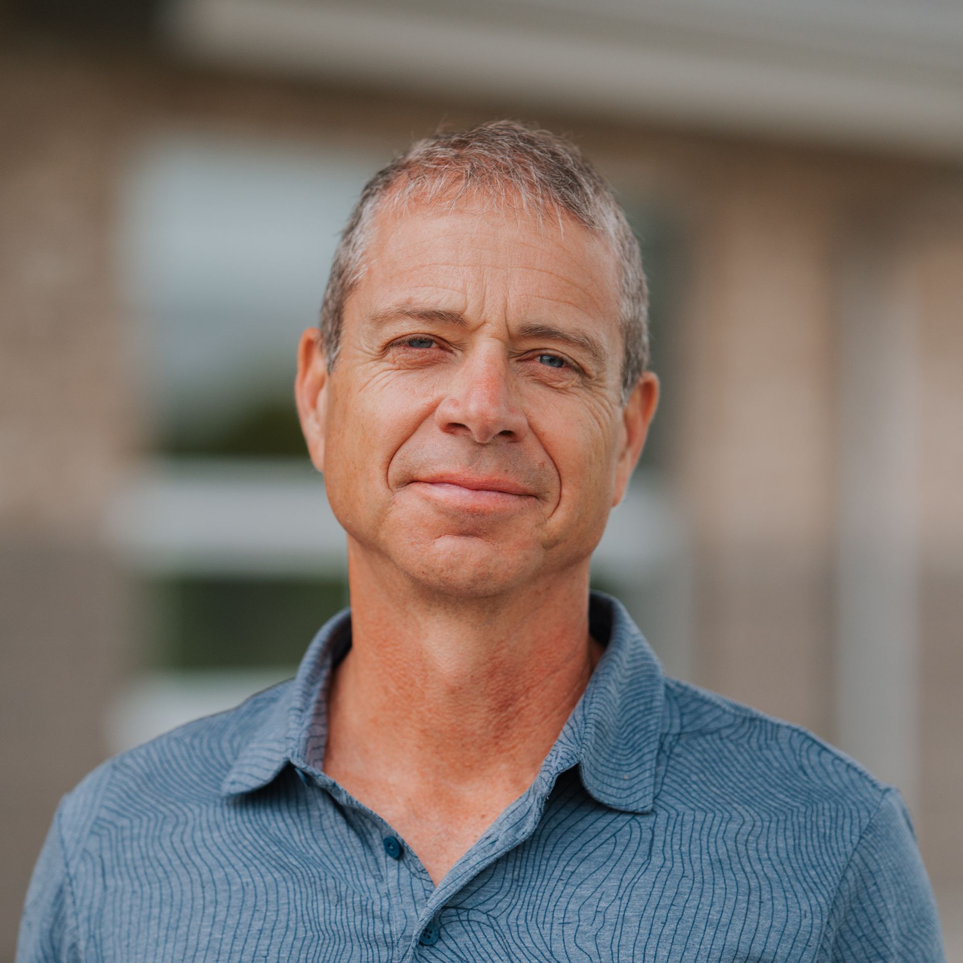 Man with short gray hair, blue shirt, looking toward the camera in front of a building with blurry windows.