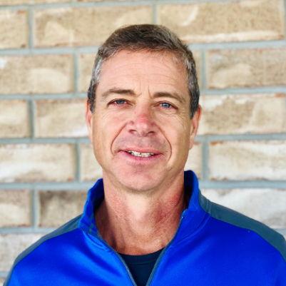 Man with graying hair in blue shirt smiling in front of a brick wall.