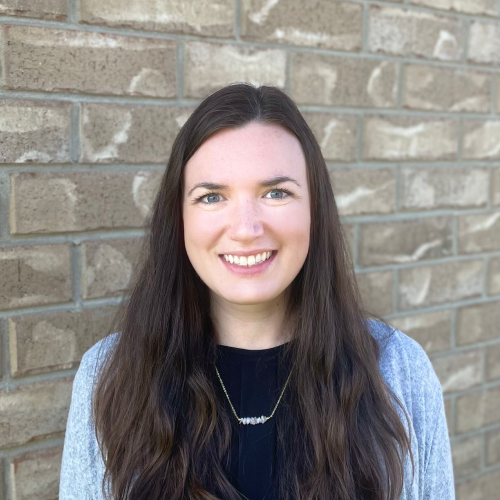 Woman with long brown hair smiles in front of a brick wall, wearing a black shirt and gray cardigan.