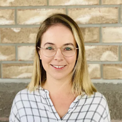 Woman with blonde hair and glasses smiles in front of a brick wall, wearing a black and white plaid shirt.