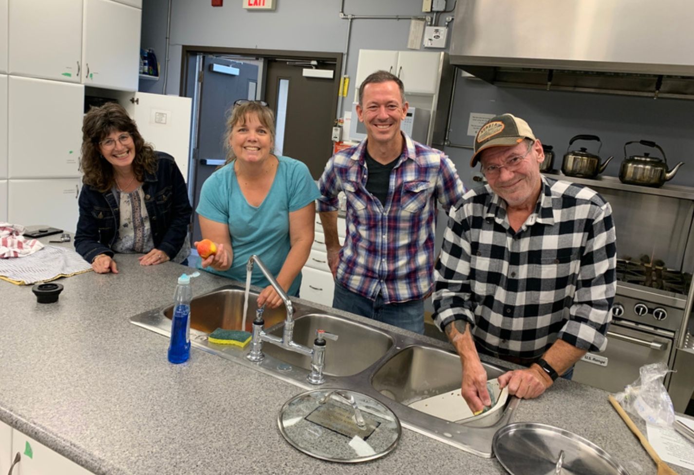 Four people in a kitchen smile. One holds a tomato, another washes dishes. Stainless steel appliances.