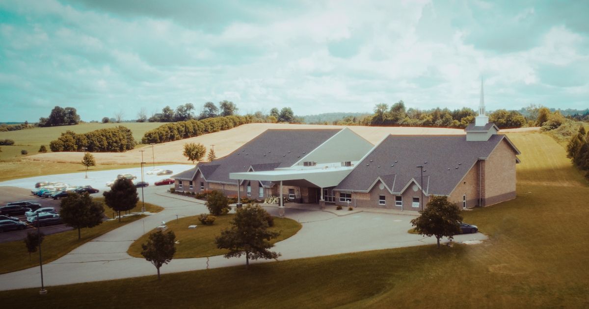 A large church building with a parking lot in a rural setting, under a cloudy sky.