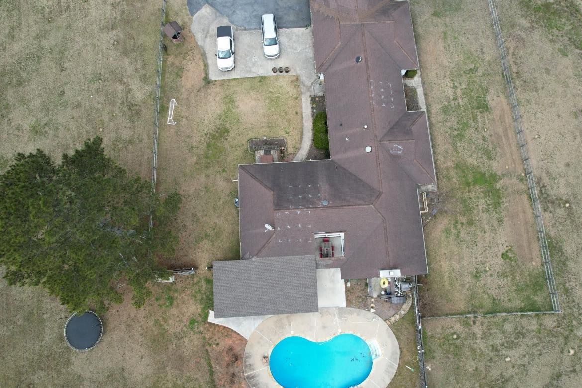 Overhead view of a house with a pool, vehicles, and a tree, surrounded by a grassy yard.