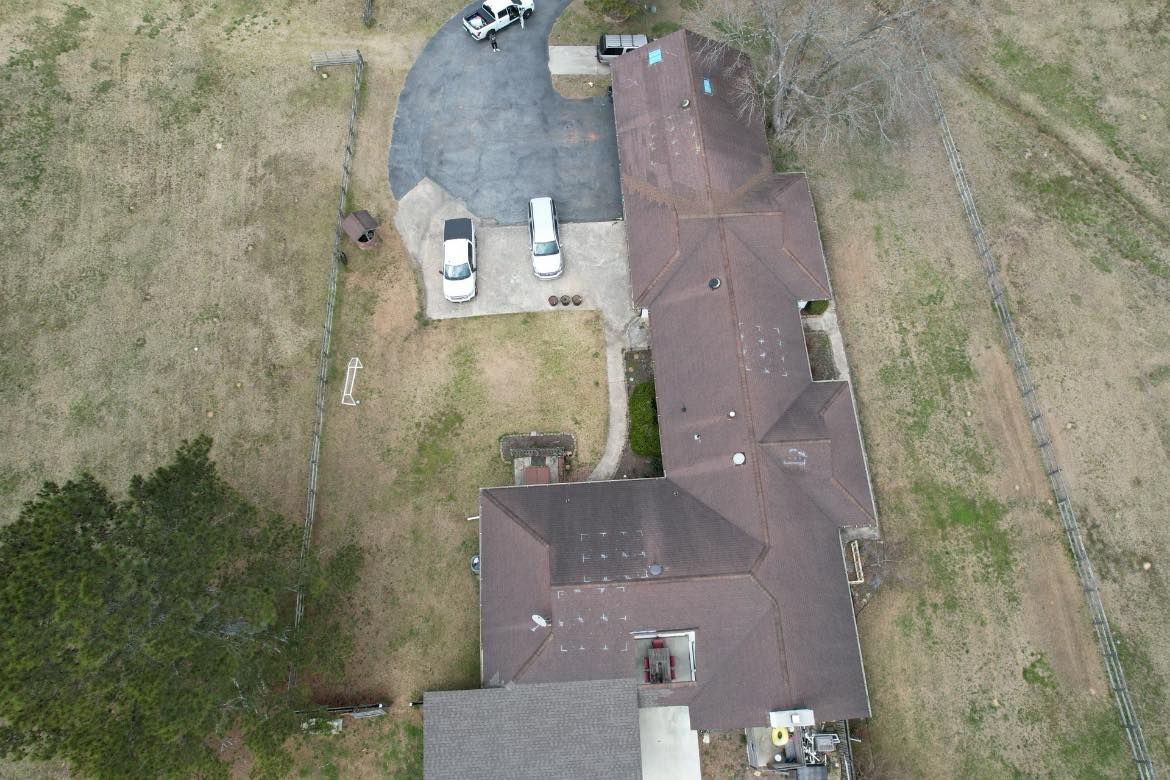 Overhead view of a brown-roofed house with a paved driveway, parked vehicles, and surrounding brown grass.