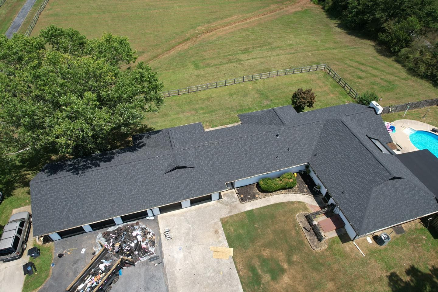 Aerial view of a house with a dark gray roof, long driveway, and pool. Dumpster on the left.