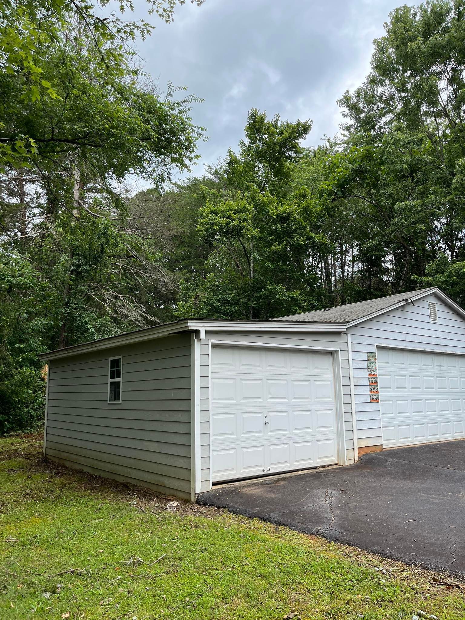 Two white garages with gray asphalt driveway, surrounded by green grass and trees under a cloudy sky.