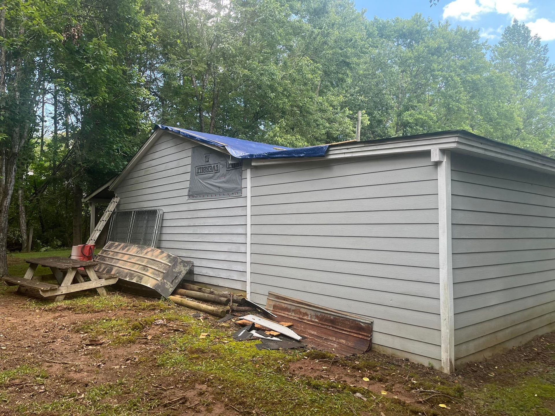 A damaged gray building with a blue tarp on the roof.  Debris and a picnic table are in the foreground.