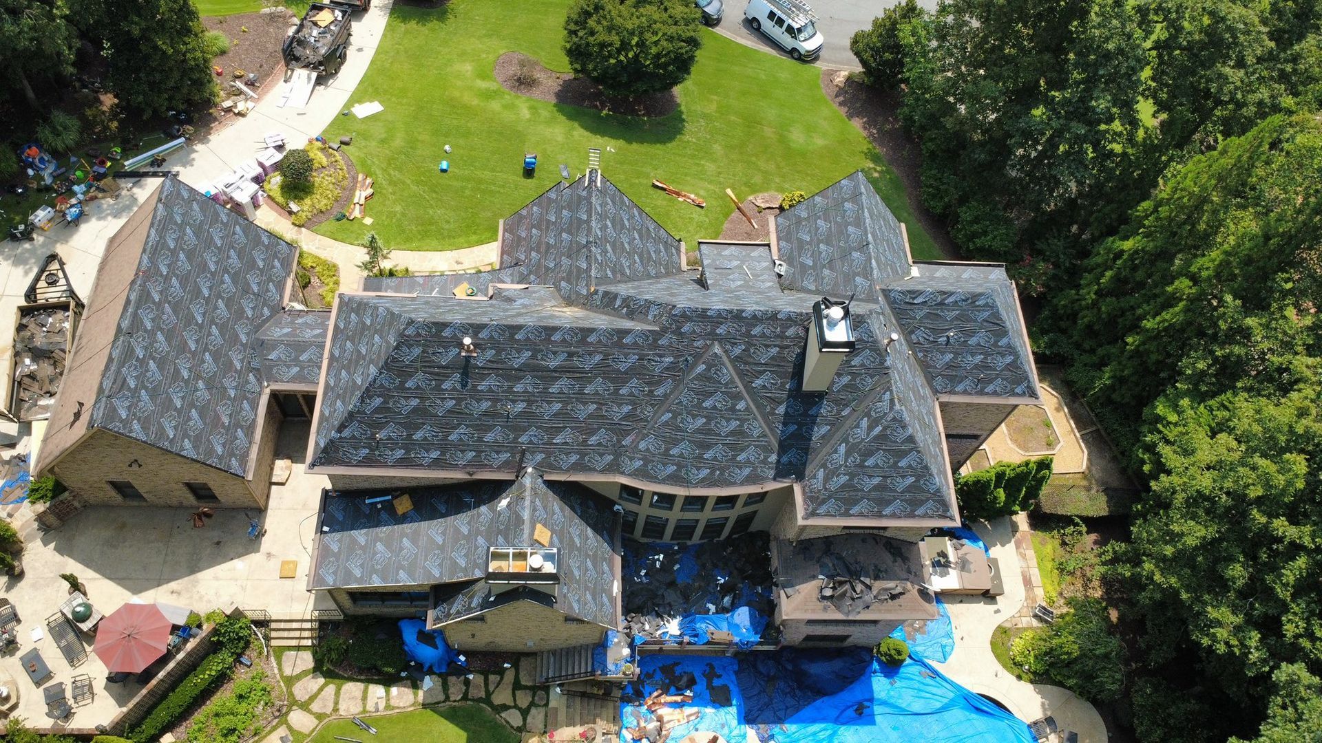 Aerial view of a large house with a dark gray roof partially covered with blue tarps, surrounded by green trees and grass.