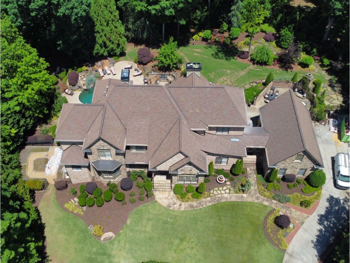 Aerial view of a large brown-roofed house with a swimming pool and landscaped yard surrounded by trees.