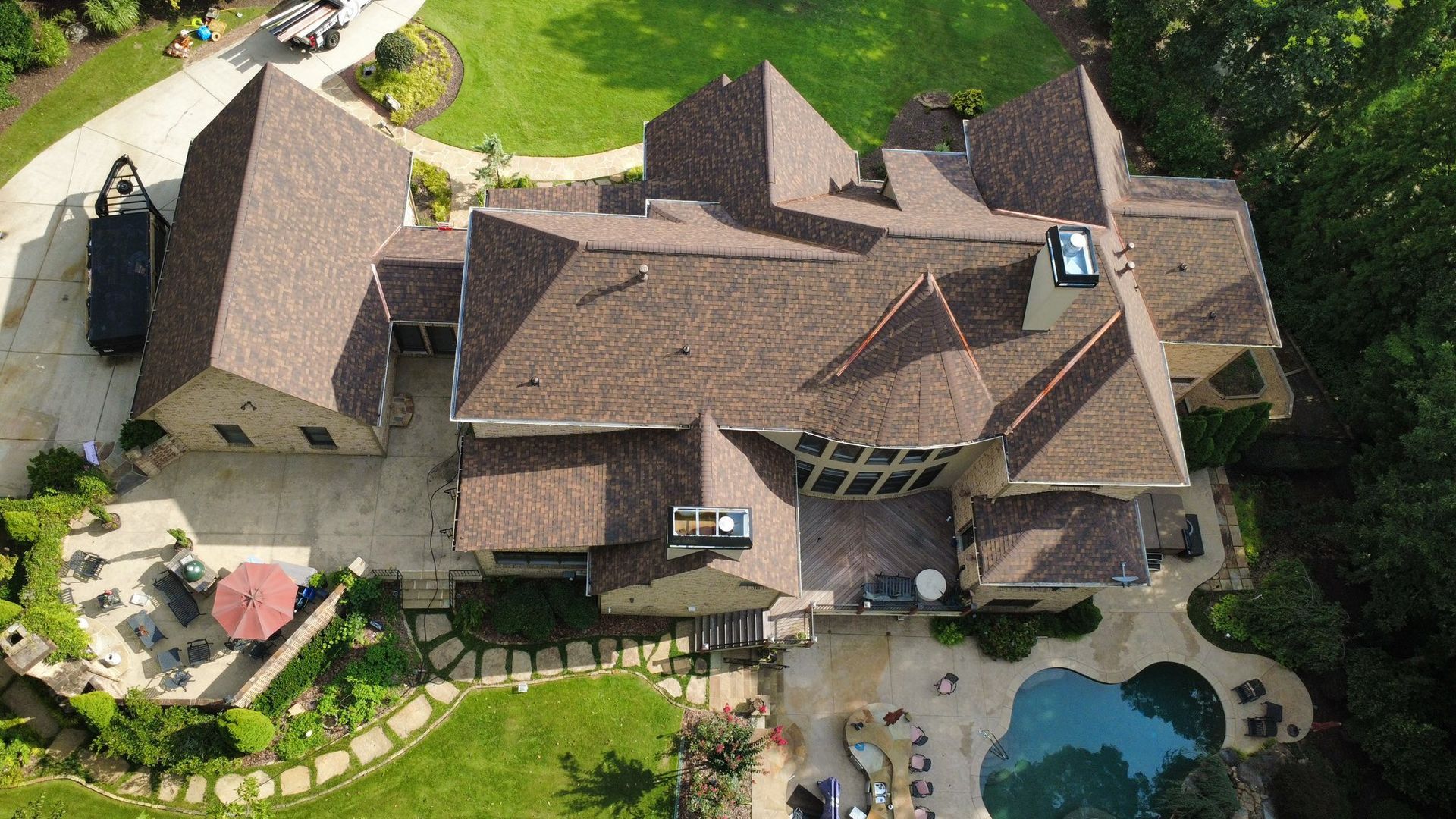 Aerial view of a large brown-roofed house with a pool, patio, and lush landscaping.