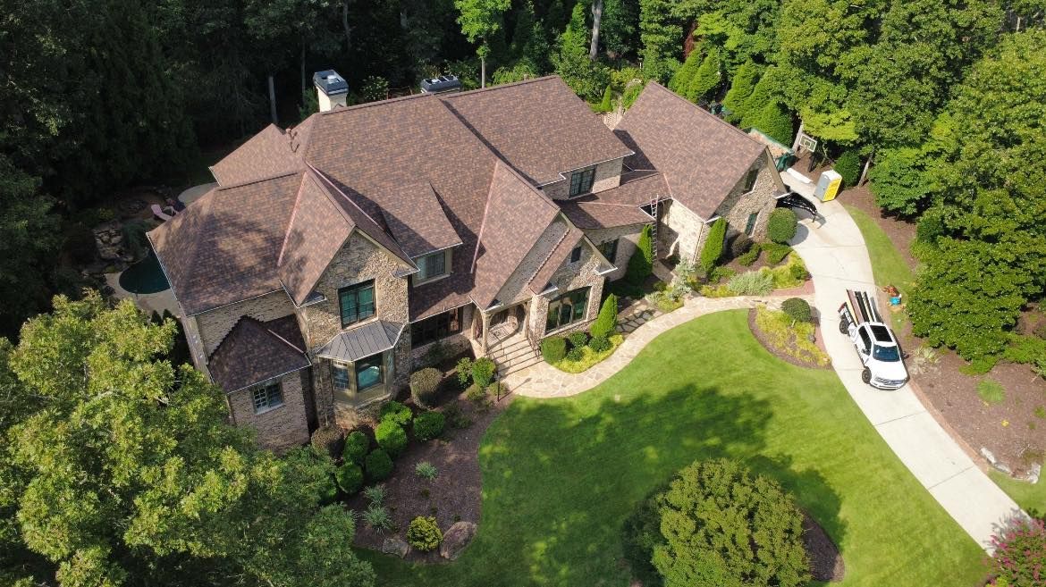 Aerial view of a large house with brown roof, surrounded by green trees and a paved driveway with a white car.