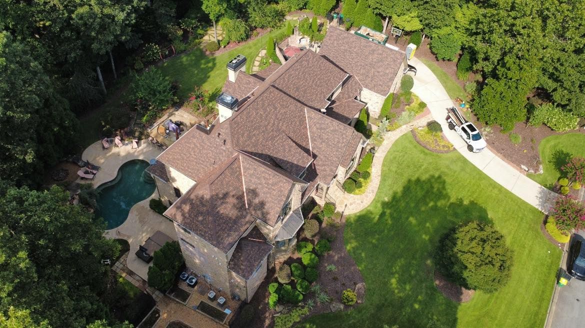 Aerial view of a large brown-roofed house with a pool, surrounded by green grass and trees.