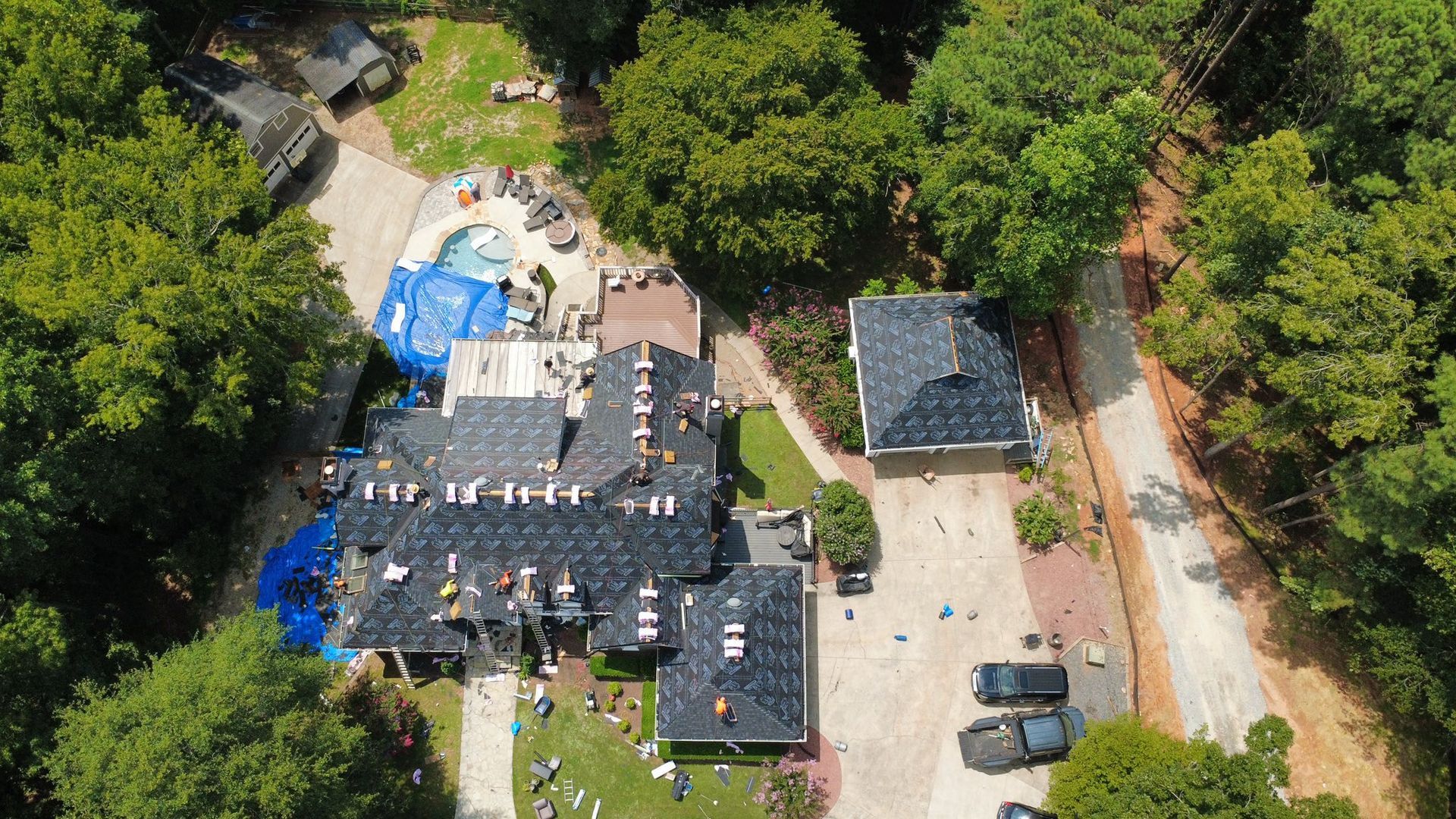 Aerial view of a house under construction; roofing being replaced; blue tarp covering a portion of the roof; surrounded by trees.