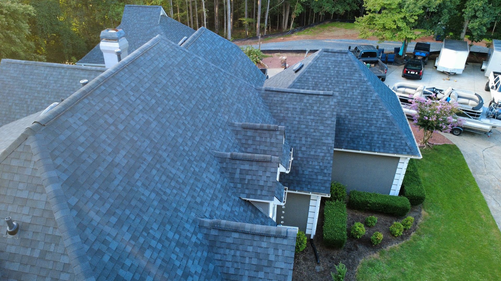Overhead view of a gray roof on a large house with a white chimney; green grass and trees in background.