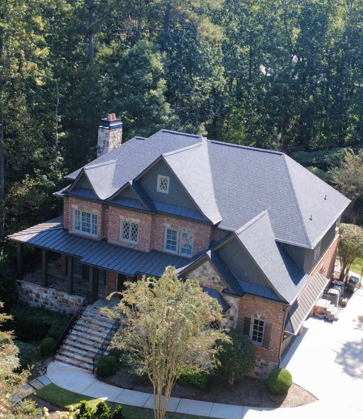 Two-story house with brick and stone exterior, dark gray roof, and a porch. Surrounded by trees and landscaping.
