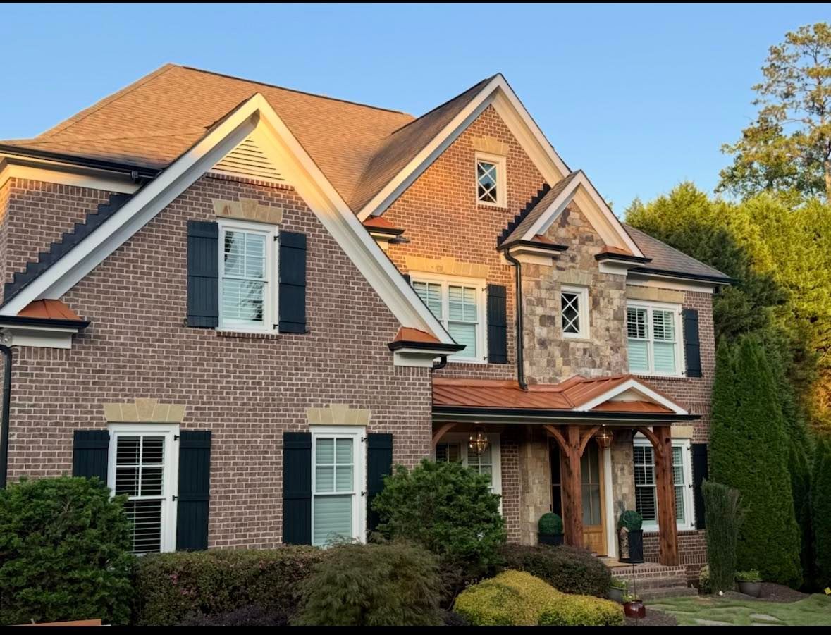 Brick and stone two-story house with black shutters, brown roof, and wooden porch under a blue sky.