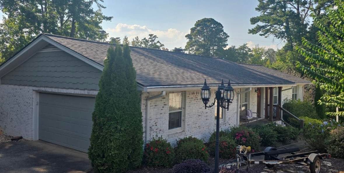 A one-story house with a garage and a brick exterior. The roof appears weathered, with overgrown shrubs in the front.