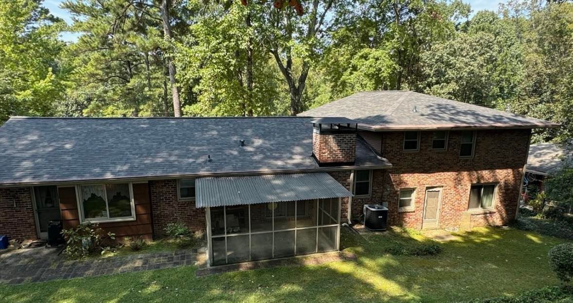 Two-story brick house with a dark roof and screened porch, surrounded by trees and green grass.