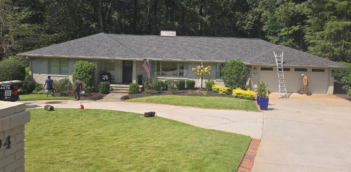 A one-story house with a gray roof, lawn, and driveway. People are working near the house.