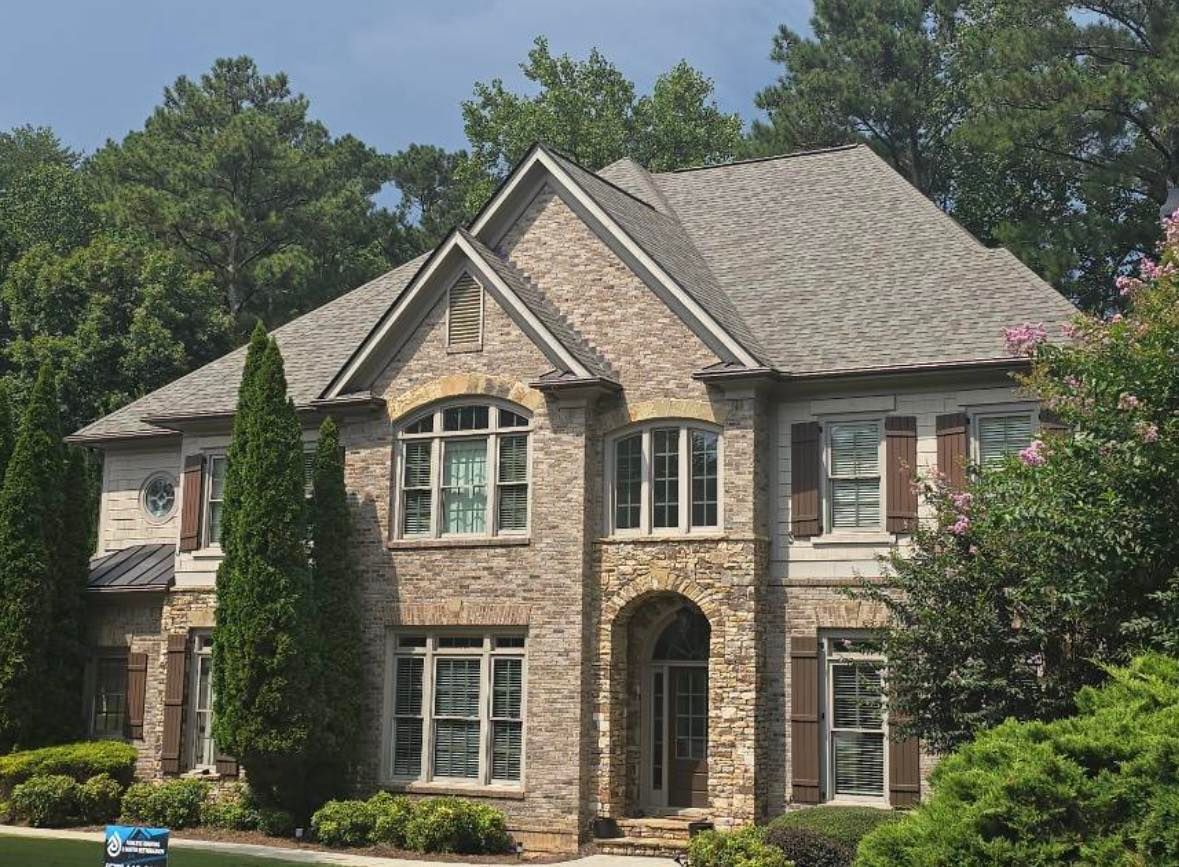 Stone-faced two-story house with arched entryway, brown shutters, and gray shingled roof, surrounded by trees.