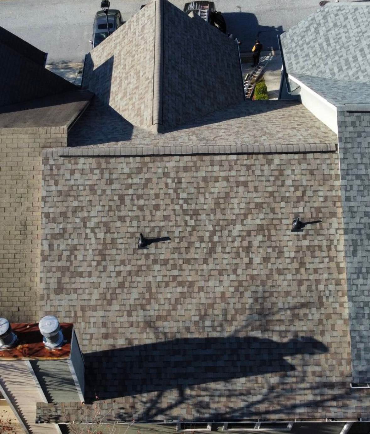 Overhead view of a house roof with brown shingles, vents, and a shadow from trees below.