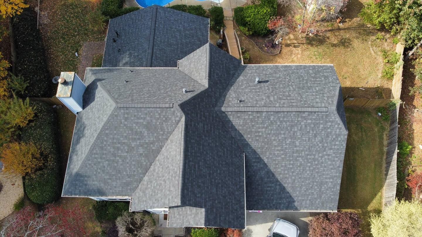 Overhead view of a house with a complex gray shingle roof, surrounded by green grass and trees.