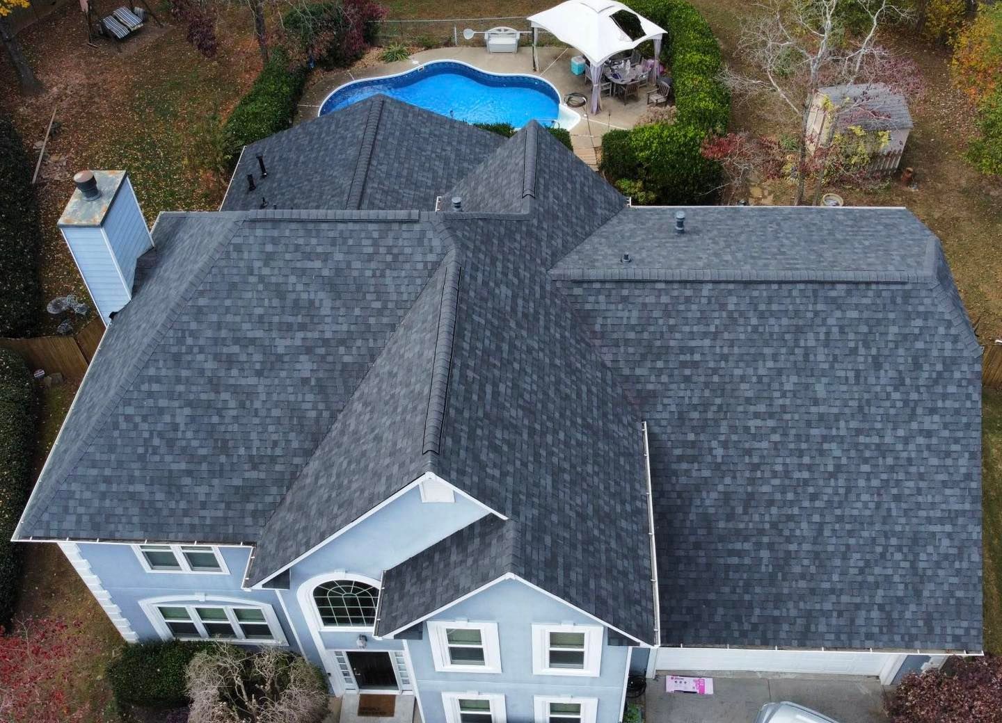 Overhead view of a blue house with a dark gray shingled roof, a swimming pool, and fall foliage.