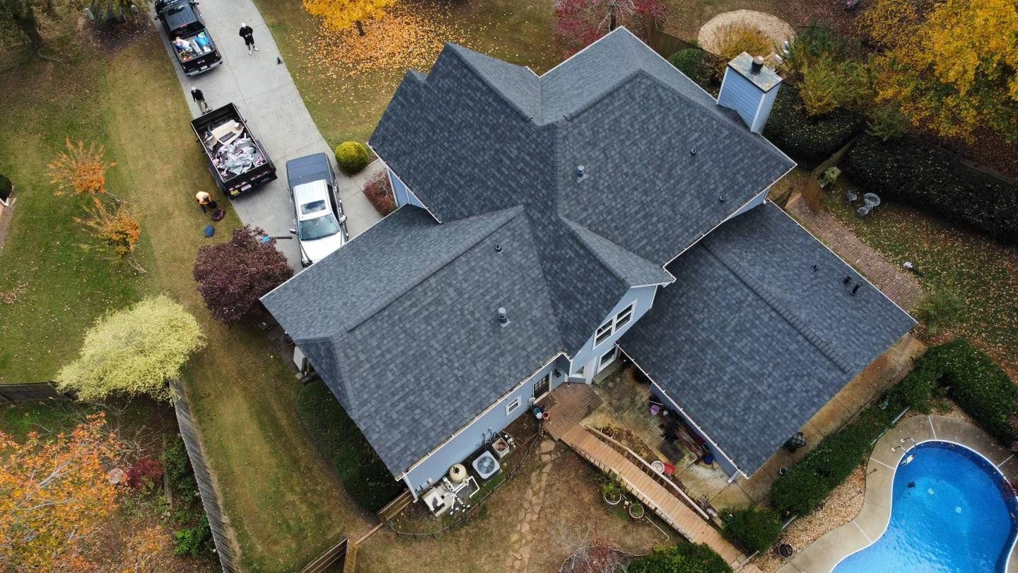 Aerial view of a gray house with a complex roof, driveway with parked vehicles, and a pool.