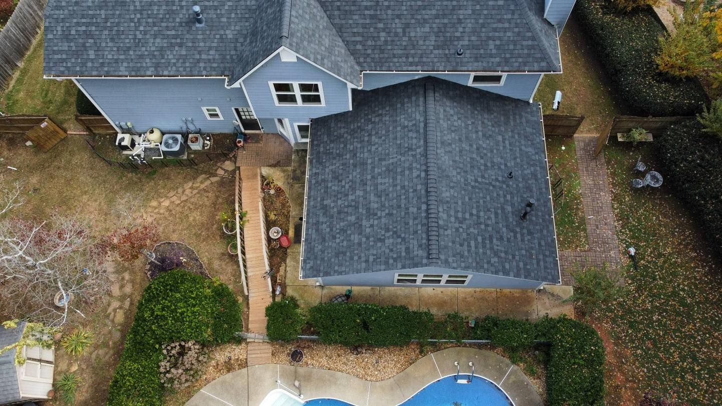 Aerial view of a blue house with a dark gray roof, a pool, and surrounding foliage.