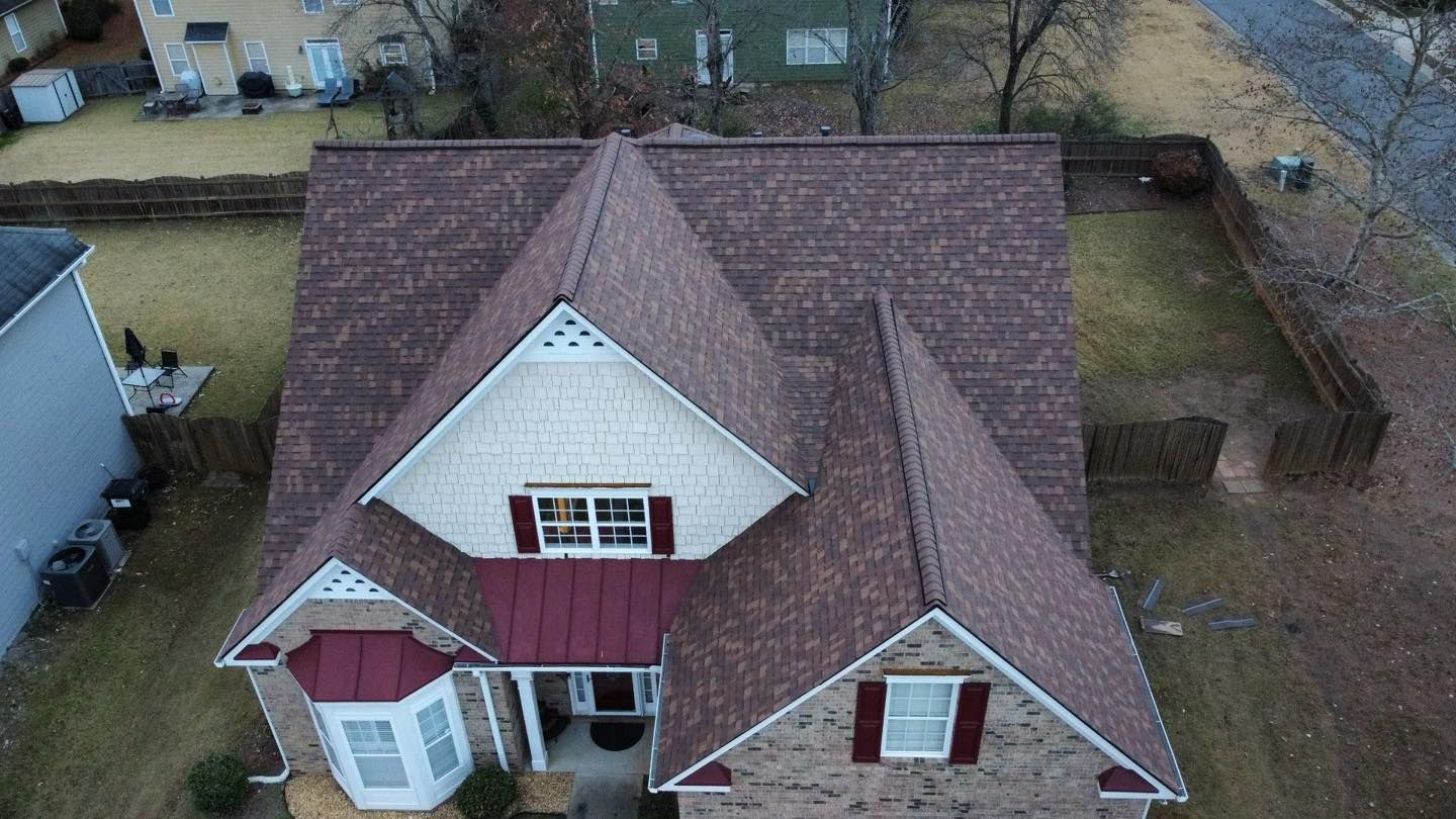 Aerial view of a brown-roofed house with white trim and red accents, surrounded by a fence and some trees.