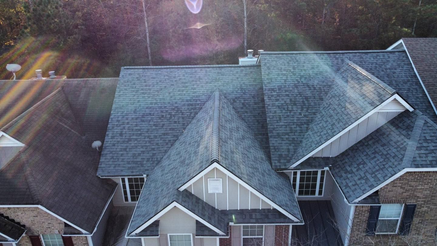 Aerial view of a gray-shingled house roof with dormers, chimney, and satellite dish. Sunlight creates a lens flare.