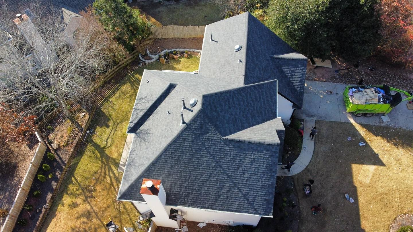 Aerial view of a house with a dark gray roof, chimney, and a green dumpster on the driveway.