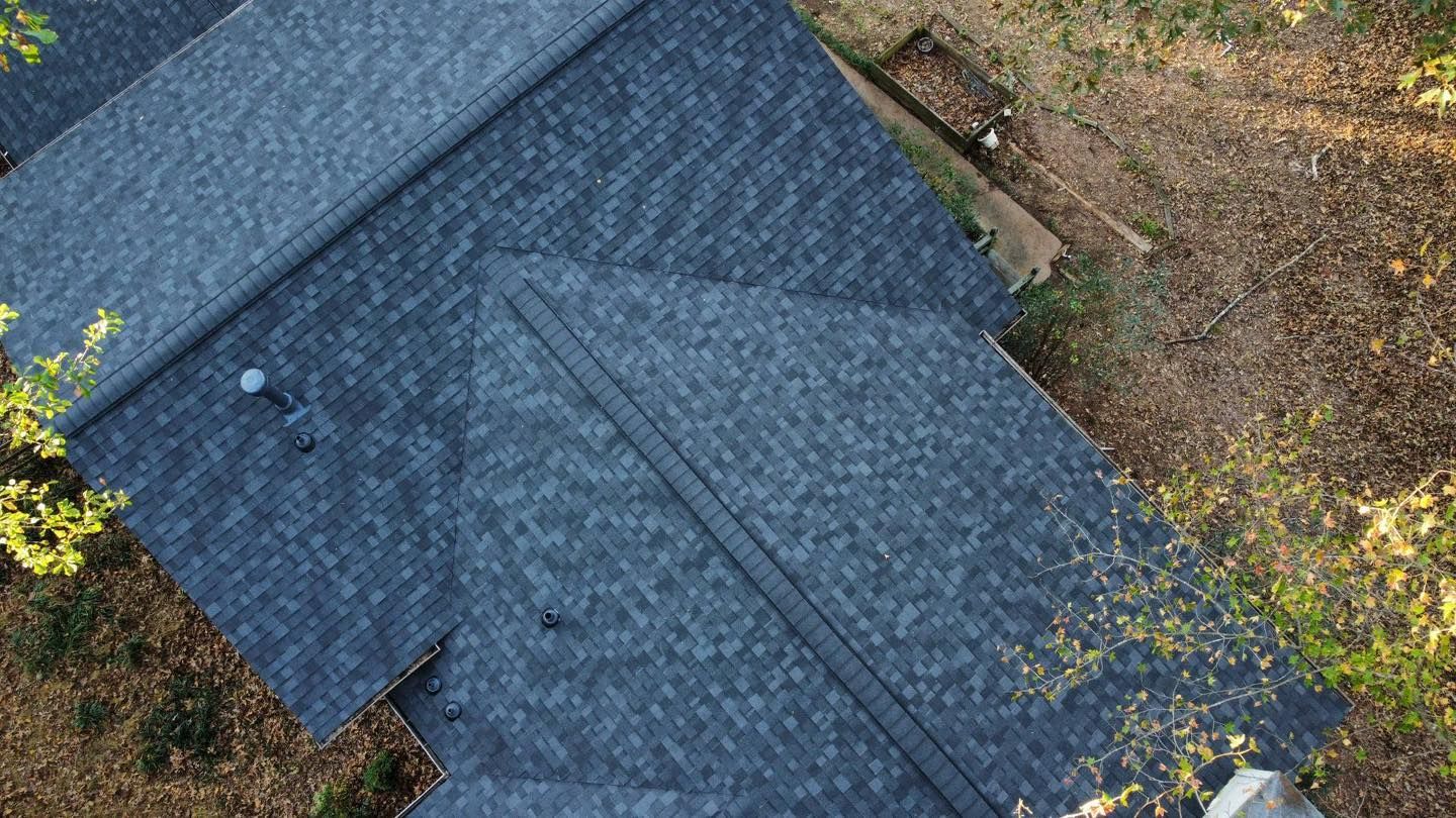 Overhead view of a house roof with dark gray shingles, surrounded by trees and foliage.