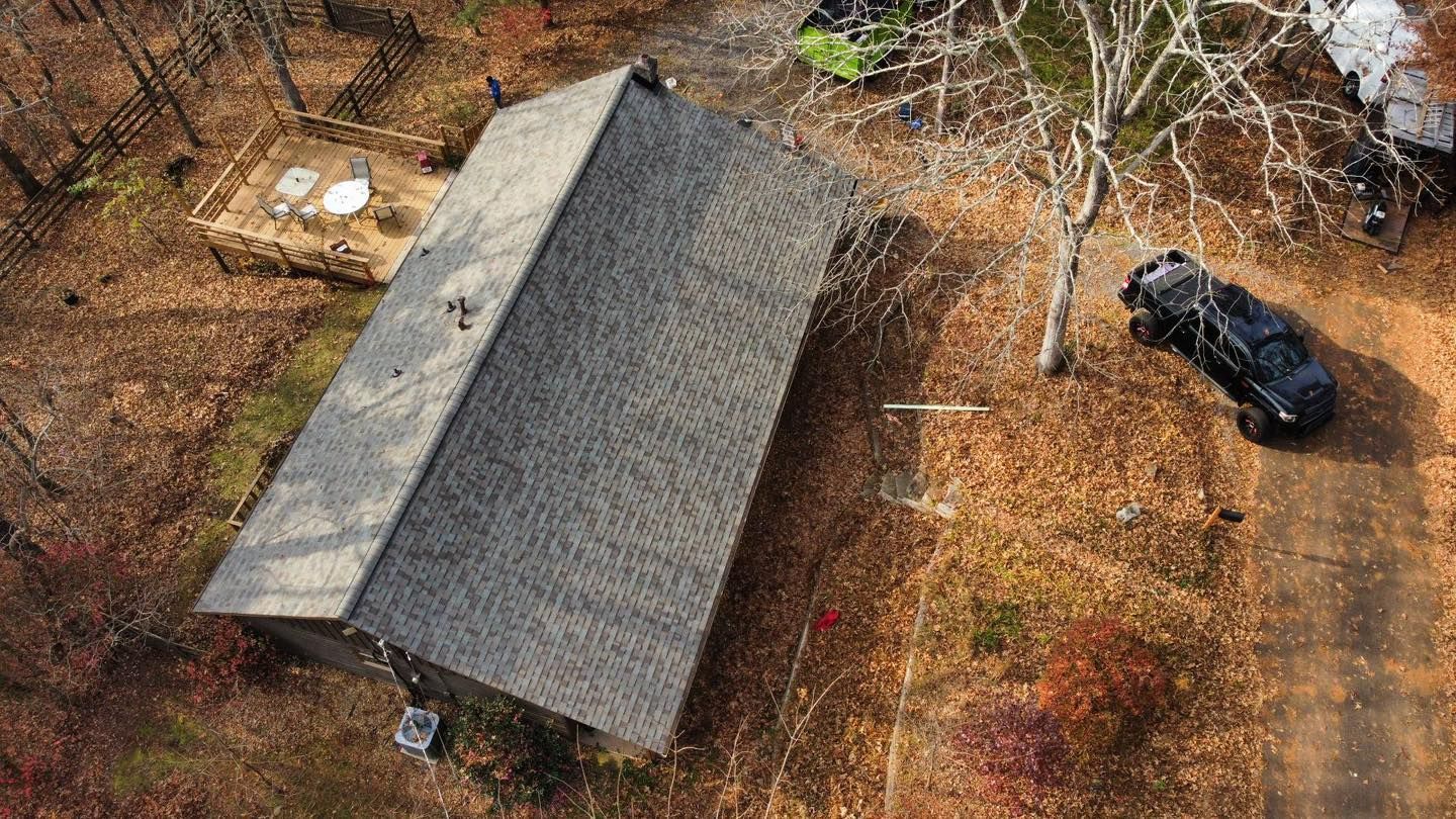Overhead view of a house with a gray roof, a black vehicle on the right, and a surrounding wooded area with fallen leaves.