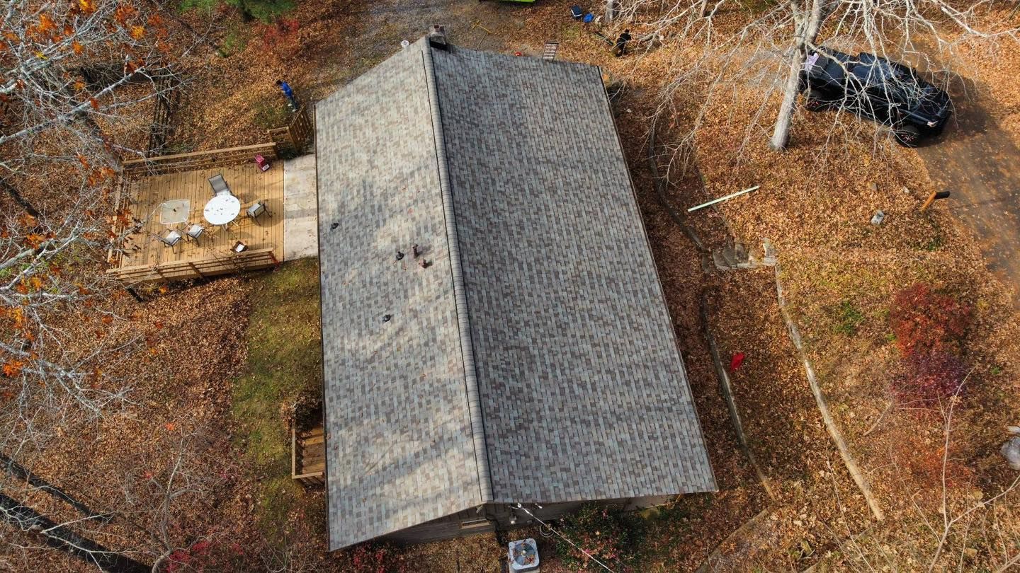 Aerial view of a house with a gray shingled roof surrounded by fall foliage, with a deck and vehicle nearby.