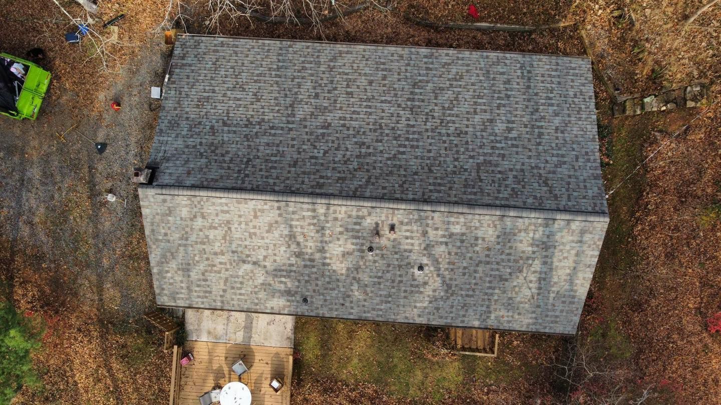 Overhead view of a house with a gray shingle roof, surrounded by brown foliage. A small patio is at the base.
