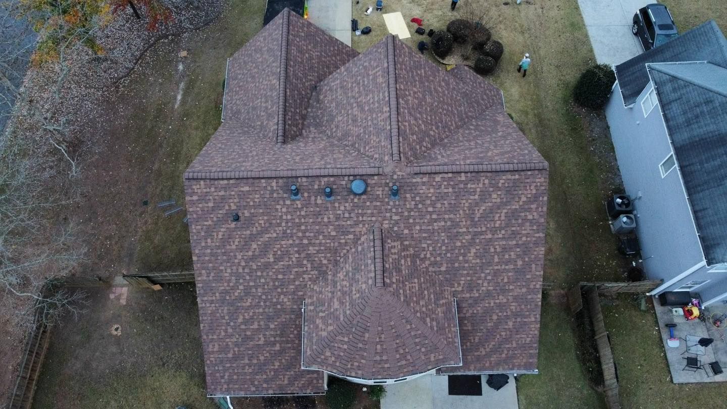 Aerial view of a brown-roofed house with two dormers, driveway, and surrounding houses in a residential area.