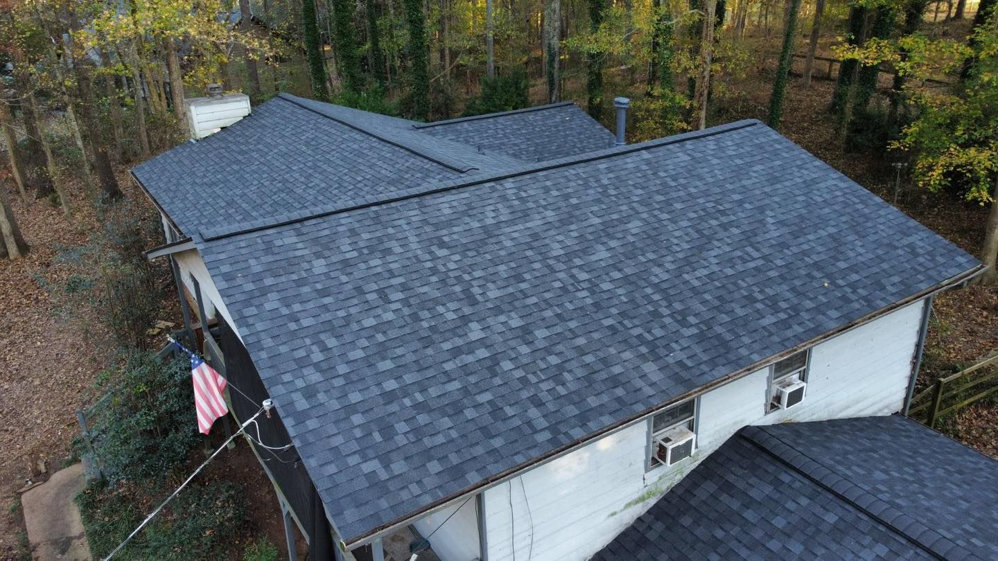 Aerial view of a white house with a dark gray shingle roof in a wooded area. An American flag hangs from the side.