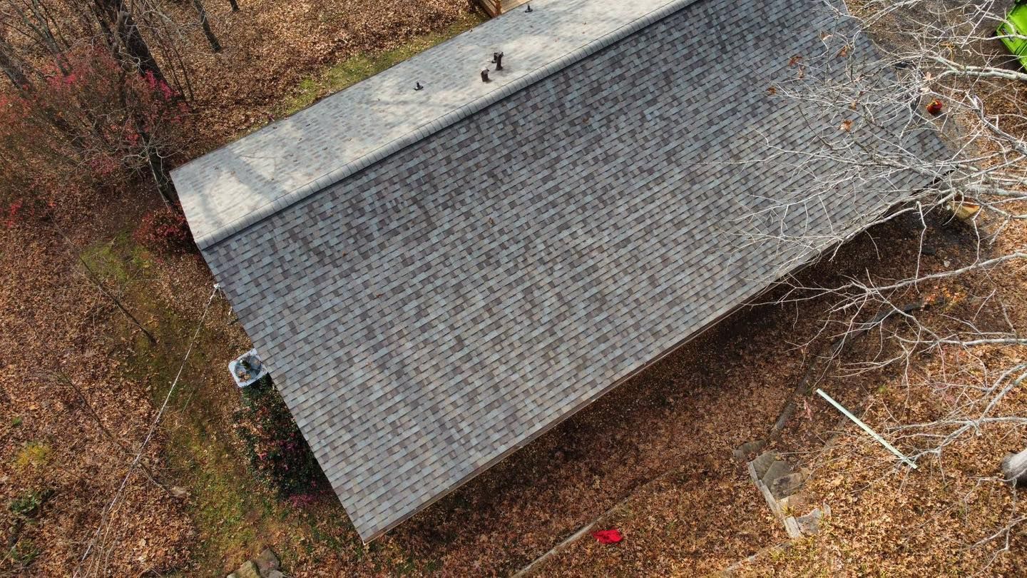 Overhead view of a gray shingled roof surrounded by brown leaves and sparse trees.